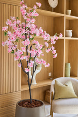 Die künstliche Kirschblüte 180 cm rosa mit ihren zartrosa Blüten steht in einem modernen Wohnzimmer mit Holzregalen, Keramik, einem beigen Sessel mit grünem Kissen und minimalistischem Dekor.