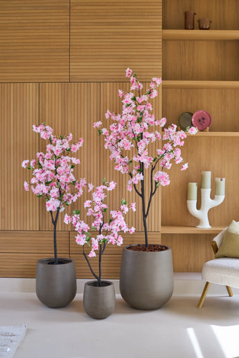 Drei 150 cm große künstliche Kirschblütenbäume mit rosa Blüten stehen vor einer Holzvertäfelung, daneben mit Dekorationen verzierte Regale und ein moderner weißer Kerzenständer.