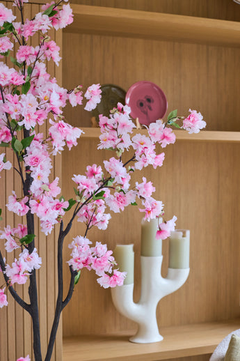 Eine künstliche Kirschblüte von 180 cm in Rosa steht vor Holzregalen, auf denen ein rosa Teller, eine grüne Schale und ein weißer Kerzenständer mit drei hellgrünen Kerzen ausgestellt sind.