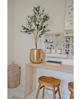Ein moderner Arbeitsplatz mit einem Holzschreibtisch, zwei Hockern, einem großen goldenen Pflanzgefäß mit dem künstlichen Olivenbaum 120 cm für mediterranes Flair, einem Waschbrett mit Büroutensilien und einem Weidenkorb auf dem Boden als dekoratives Detail.