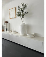 Ein minimalistischer Raum mit einem weißen Sideboard, dem künstlichen Olivenbaum 120 cm als Dekoelement, dekorativen weißen Schalen, einer Kerze und abstrakter Wandkunst. Dunkle Holzböden und moderne Akzente schaffen eine ruhige mediterrane Atmosphäre.