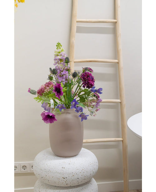 Eine künstliche Blumen-Glockenblume von 62 cm Lila steht in einer Milchglasvase auf einem strukturierten weißen Sockel. Dahinter lehnt eine helle Holzleiter an einer beigen Wand, in Bodennähe sind Steckdosen sichtbar.