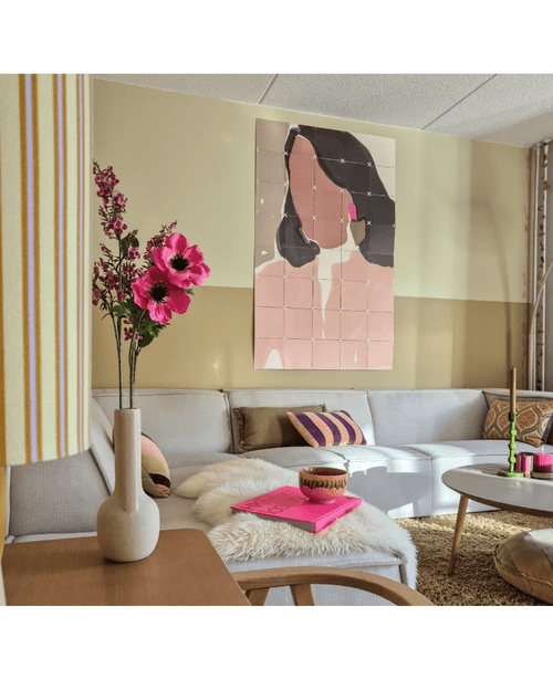 Ein gemütliches Wohnzimmer mit einem hellgrauen Sofa, rosa und goldenen Akzenten, einer 56 cm großen künstlichen Anemone in rosa Zweigen in einer modernen Vase, einem Couchtisch mit einem Fellteppich und abstrakter Pastellkunst einer Frau an der Wand.