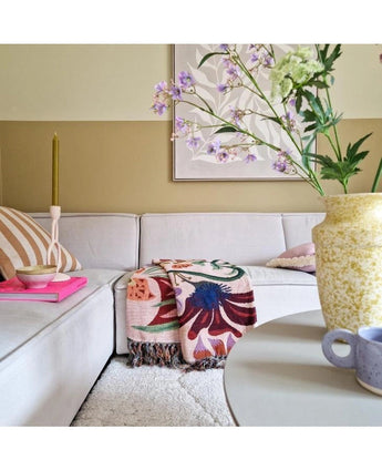 Ein gemütliches Wohnzimmer mit einem hellbeigen Sofa, einem geblümten Plaid, einer rosa Kerze, einem gestreiften Kissen und einer künstlichen Spitzenblume 76 cm weiß in einer gelben Vase auf dem runden Tisch. Abstrakte Kunst schmückt die Wand dahinter.