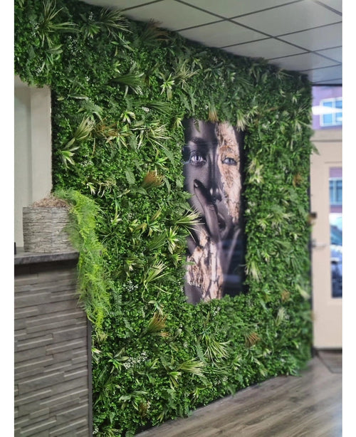 Eine mit künstlicher Heckenmatte Savanne 100x100 cm bedeckte Wand, mit UV-geschütztem Grün in einem Kunststoffpflanzgefäß Ø100, hat in der Mitte ein großes schwarz-weißes Porträt. Der moderne Innenraum verfügt über eine geflieste Decke und eine Holztheke auf der linken Seite.