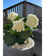 Die künstliche Hortensie White UV 35 cm mit weißen Blüten und grünen Blättern steht an einem sonnigen Tag draußen auf weißen Kieselsteinen, mit Terrassengeländer aus Holz und einer Treppe im Hintergrund.