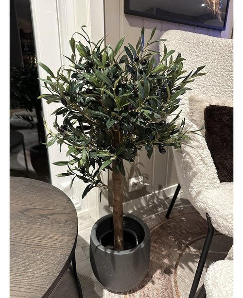 Der künstliche Olivenbaum UV 85 cm, eine kompakte und pflegeleichte Kunstpflanze mit dunkelgrünen Blättern und schwarzen Oliven, steht drinnen neben einem runden Holztisch und einem Sessel mit weißer Struktur.