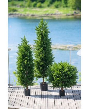 Drei künstliche Pflanzenkoniferen Deluxe UV 120 cm schmücken eine Holzterrasse mit Glasgeländer und Blick auf Wasser und Grün – dank ihres UV-Schutzes ideal für jeden Kunstpflanzengarten.