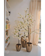 Eine 120 cm hohe künstliche Bougainvillea-Pflanze mit weißen Blüten in einem strukturierten Goldtopf steht neben einem Fenster mit transparenten Vorhängen, neben Regalen voller Bücher und Dekoration in einem hellen, modernen Raum.