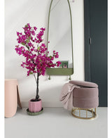 Ein modernes Interieur mit der künstlichen Bougainvillea 125 cm Lila in einem dekorativen Topf, einem hohen Spiegel mit grünem Rahmen, einem rosa Fußhocker mit goldenem Sockel und rosa Decke und einer dunklen Tür vor einer strahlend weißen Wand.