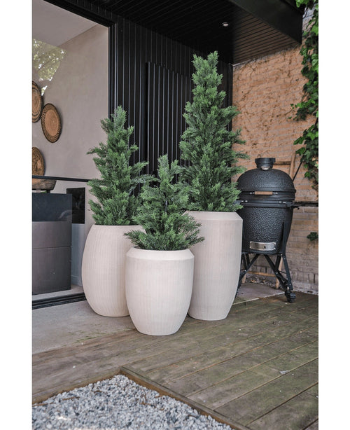Auf einer Holzterrasse neben einem modernen Gebäude stehen drei hohe, weiße Pflanzgefäße mit UV-künstlichem Nadelbaum (90 cm). Ein schwarzer Keramikgrill steht in der Nähe vor einer mit Weinreben gesäumten Steinmauer.