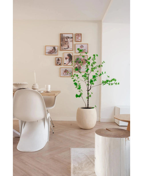 Ein moderner, minimalistischer Essbereich mit weißen Stühlen, einem Holztisch und der 150 cm großen künstlichen Enkianthus-Pflanze, die für Grün sorgt, ergänzt durch eine Galeriewand mit gerahmten Fotos auf einem neutralen Hintergrund.