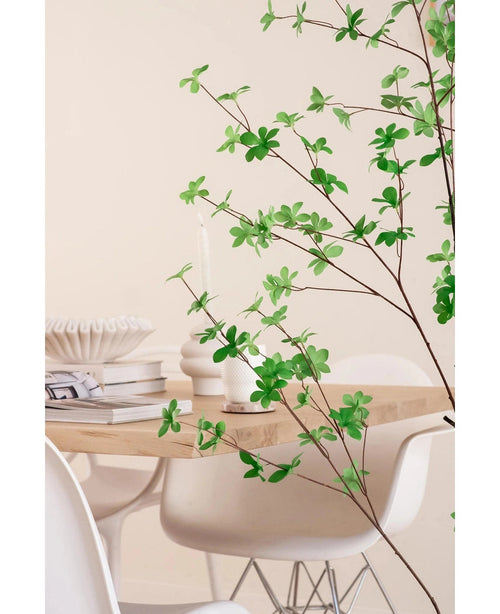 Ein moderner Essbereich mit weißen Stühlen und einem hellen Holztisch, dekoriert mit Büchern, Kerzen und einer weißen Vase, wird im Vordergrund von der künstlichen grünen Blattpflanze Enkianthus 150 cm eingerahmt.