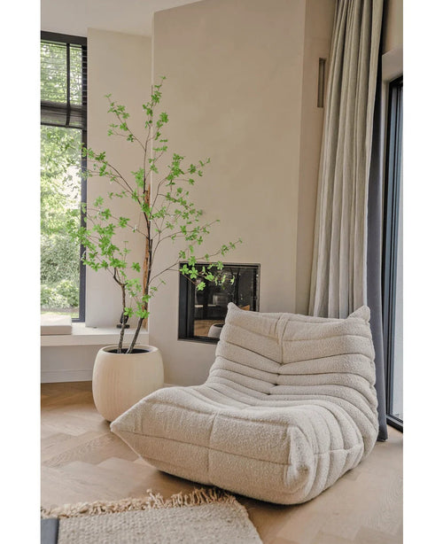 Ein gemütlicher, cremefarbener Struktursessel steht neben der künstlichen Pflanze Enkianthus 210 cm und einem modernen Kamin in einem hellen, minimalistischen Wohnzimmer mit hellen Holzböden und großen Fenstern.
