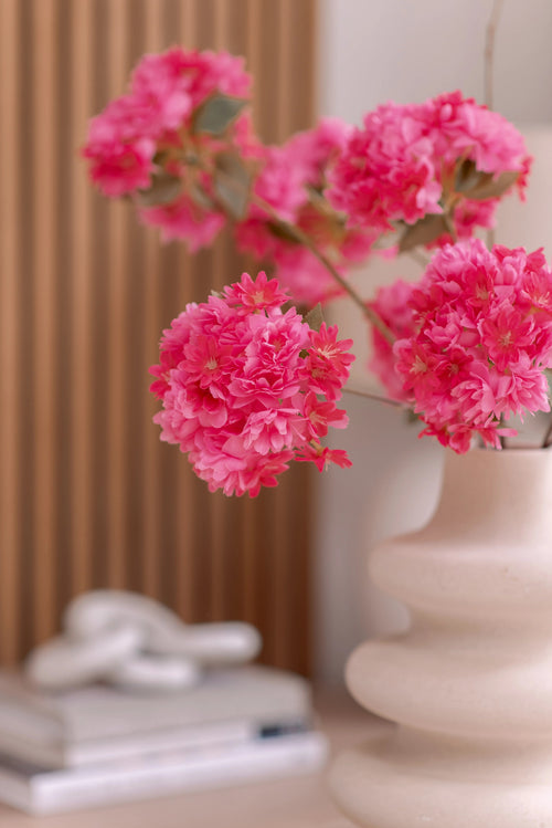 Eine weiße Keramikvase enthält einen künstlichen Kirschblütenzweig 85 cm rosa. Der Hintergrund zeigt vertikale Holztafeln, einen Stapel Bücher und darauf ein kleines Dekorationsobjekt.