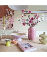 Auf einem hellen Holztisch steht die kleinblumige Art Branch Magnolia (89 cm) weiß in einer rosafarbenen Vase, moderne Keramik, bunte Tassen, eine Schale, eine gelbe Kerze in einem einzigartigen Halter und ein rosafarbenes Buch, an der Wand dahinter hängen Blumenkunstwerke.