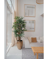 In einem hellen, modernen Wohnzimmer mit weißem Sofa, beigen Kissen, geflochtenem Tisch und abstrakter Kunst steht eine 180 cm große künstliche Palme in der Nähe eines großen Fensters. Mit seinen neutralen Farbtönen und dem Charme grüner Kunstpflanzen sorgt es für einen gemütlichen, minimalistischen Stil.