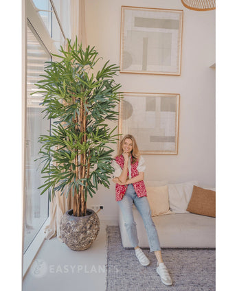 Eine Frau in rosa gemusterter Strickjacke und Jeans lächelt auf einem beigen Sofa neben der Lady Artificial Palm 210 cm in einem hellen, modernen Wohnzimmer mit abstrakter Kunst an der Wand.