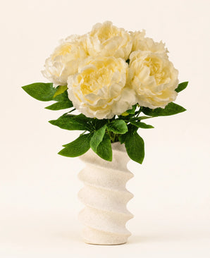 In einer gewellten, strukturierten weißen Vase befindet sich der Seiden-Kunstblumenstrauß Peony White – große cremefarbene künstliche Pfingstrosenblüten mit grünen Blättern vor einem schlichten hellen Hintergrund.