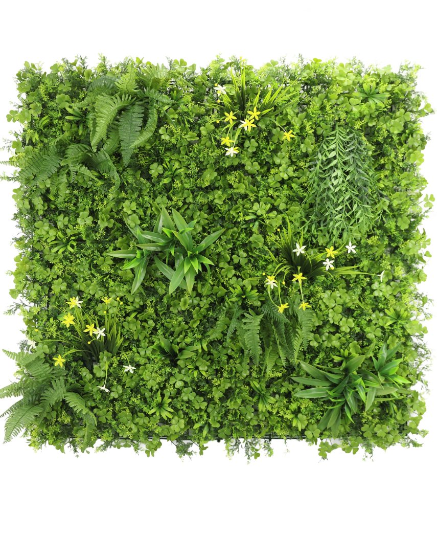 Die künstliche Hecke Narzisse Smile 100x100 cm UV ist eine quadratische vertikale grüne Wand mit dichtem grünem Blattwerk, Farnen und kleinen gelben und weißen Blüten vor einem weißen Hintergrund, von oben gesehen.